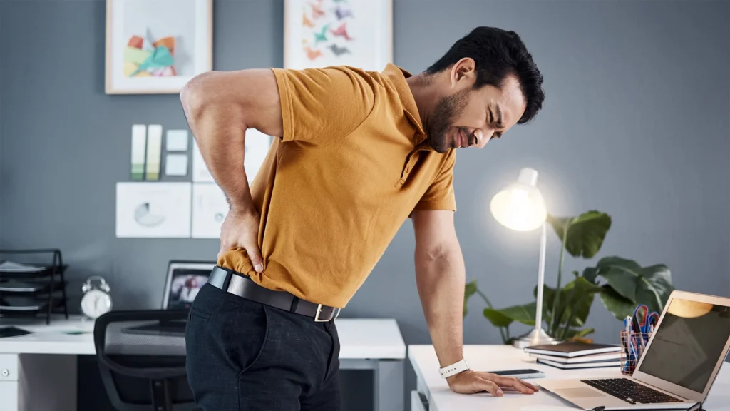 man leans on office desk as he clutches his lower back in pain